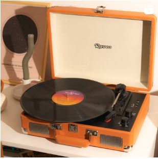 A imagem apresenta uma vitrola portátil Raveo em uma maleta laranja, com um disco de vinil pronto para tocar. Ao fundo, há um objeto decorativo moderno e a capa de um disco, sugerindo um ambiente acolhedor e musical.